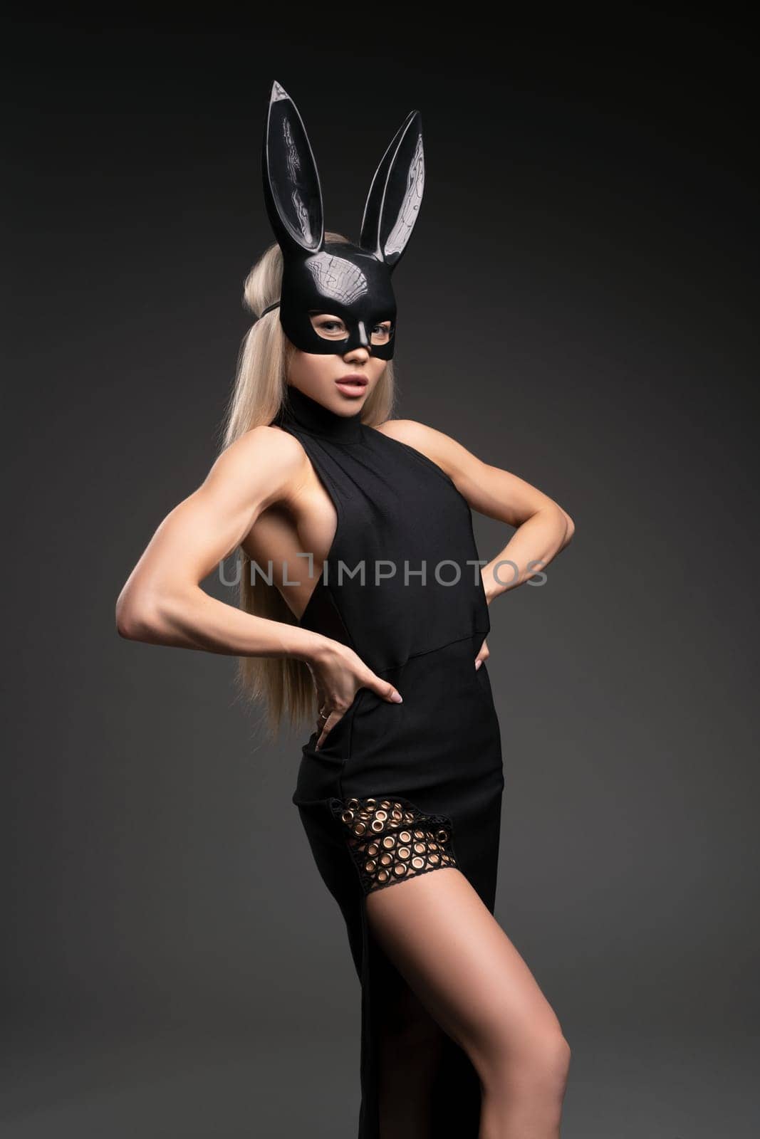 Alluring young fit female model with long blond hair in trendy black dress and latex bunny mask standing looking at camera