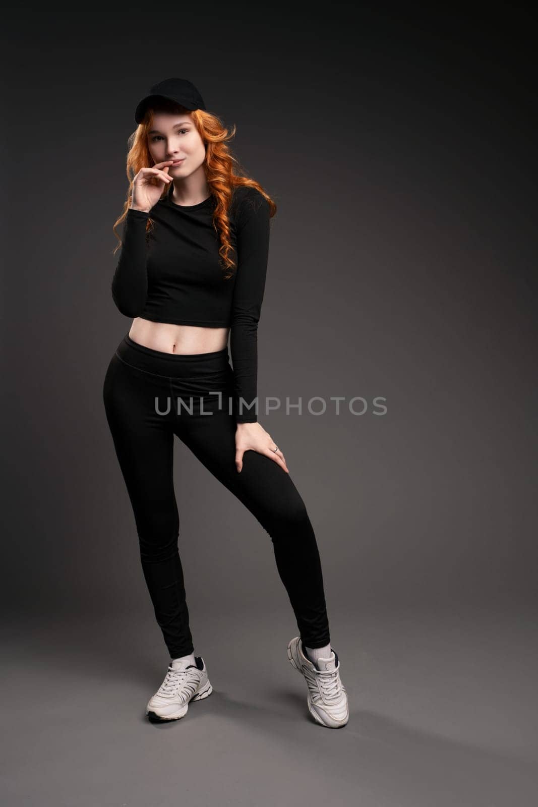 Self assured young female model with curly long red hair in trendy activewear adjusting cap and looking at camera while leaning forward against gray background