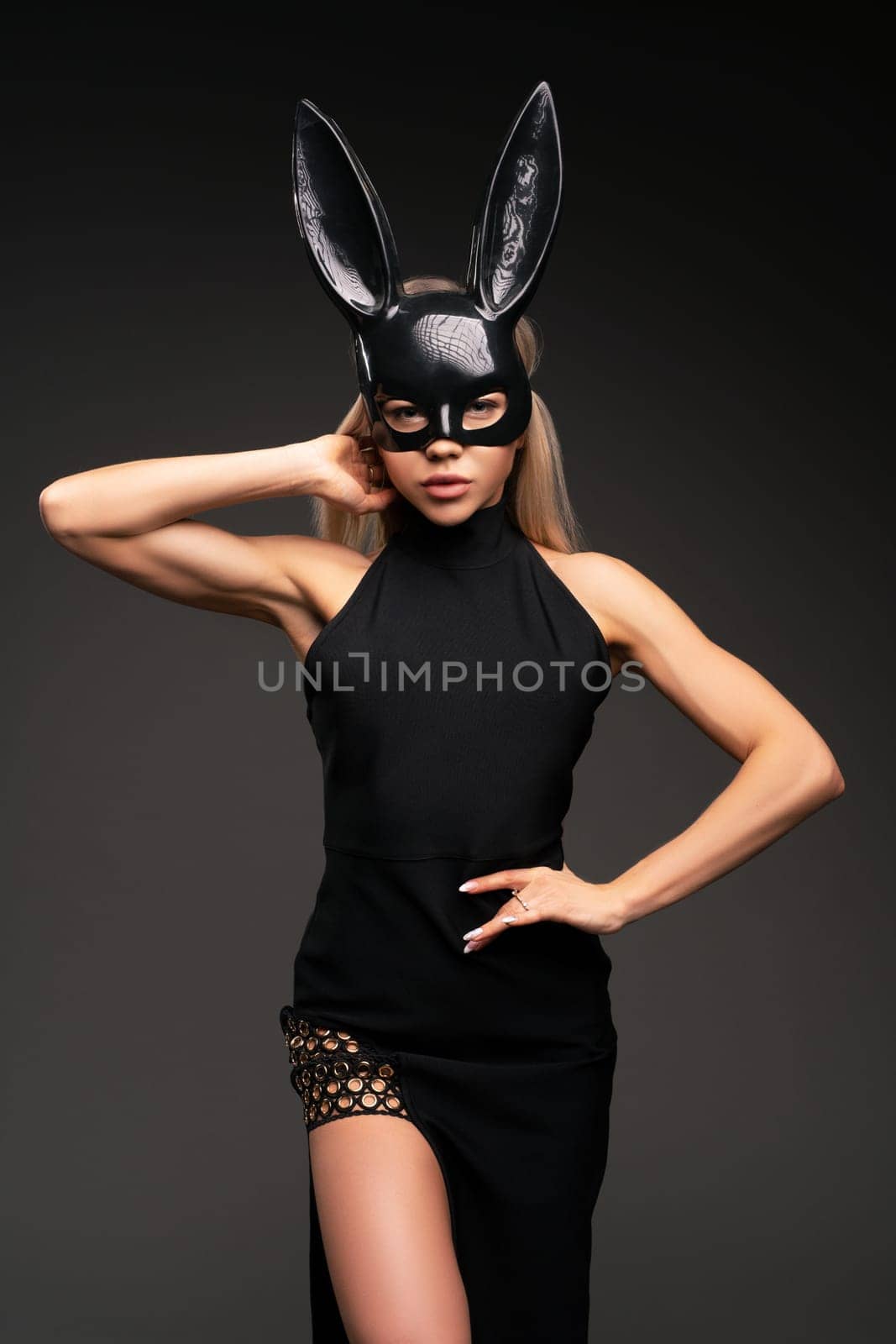 Portrait of sexy young fit female model with long blond hair in trendy black dress and latex bunny mask look at camera with seduction