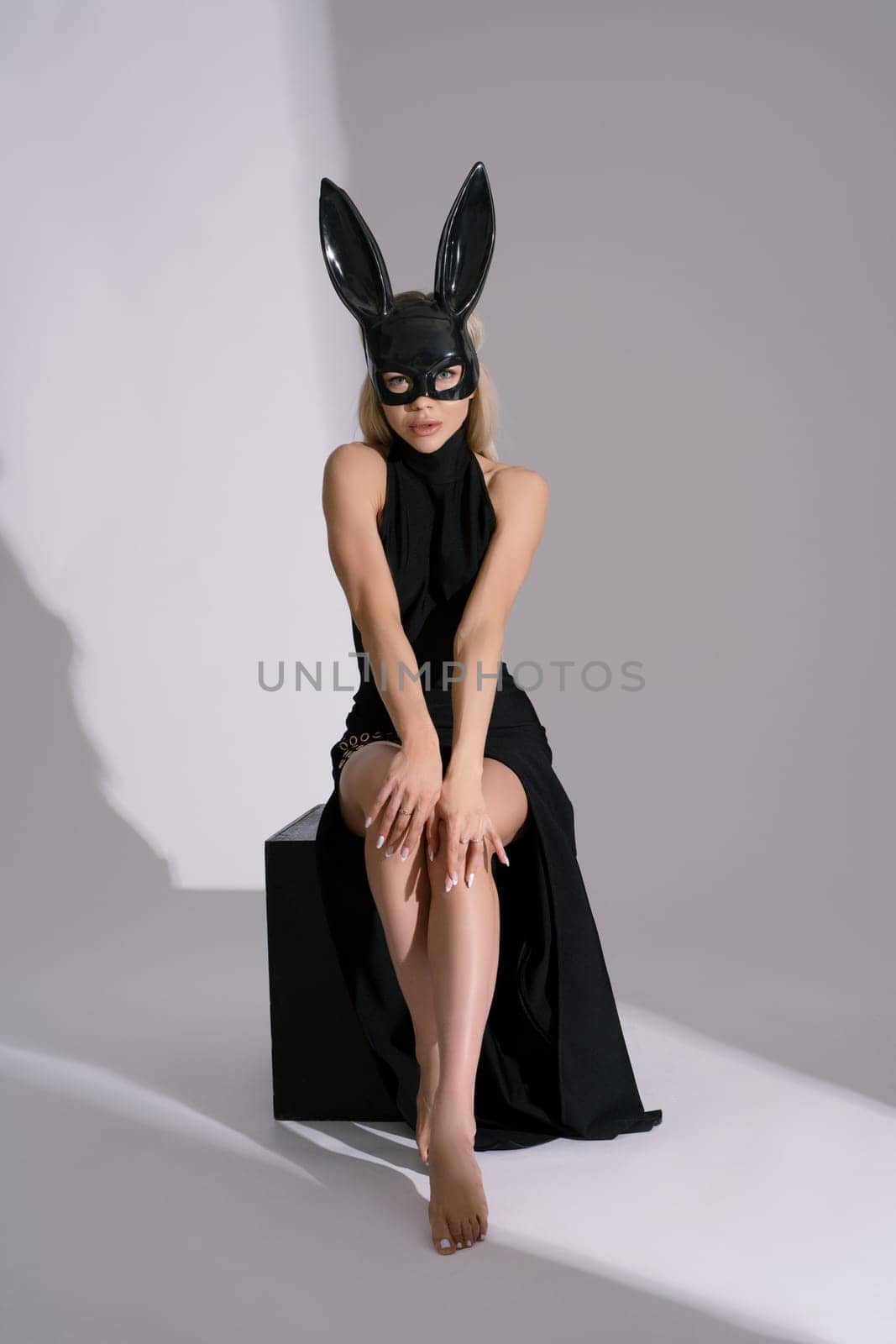 Full body of self assured barefooted young female model in black dress and rabbit mask sitting on cube - looking at camera against white background