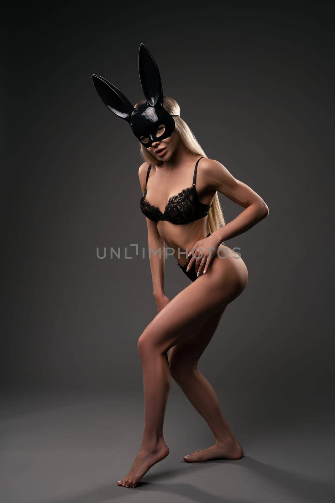 Full body of sensual young fit female model with long blond hair in sexy black lingerie and rabbit mask standing against gray background