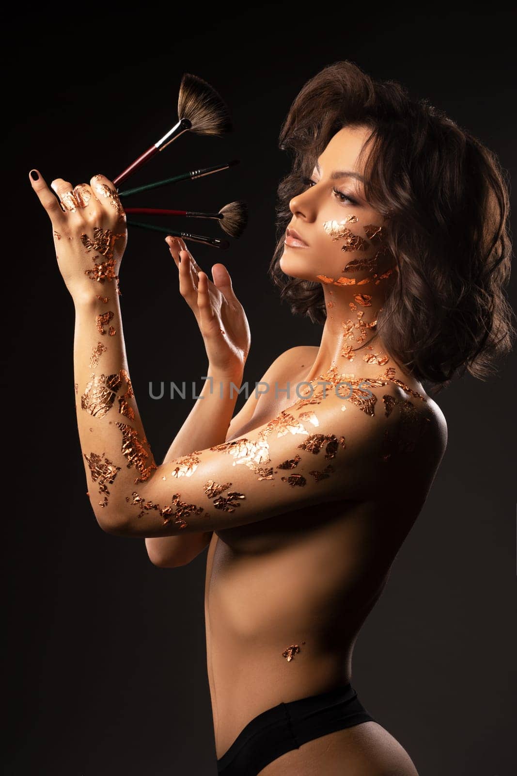 Side view of sensual young female model in black lingerie touching face and makeup brush while standing against black background
