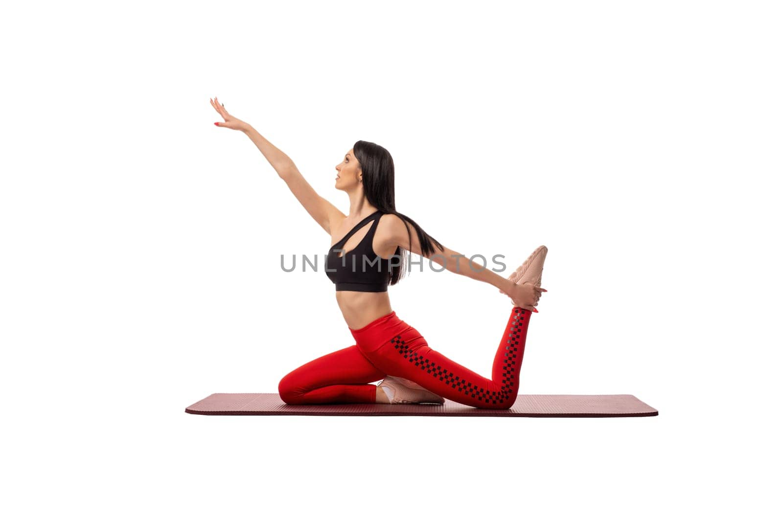Slim female in red leggings and black crop top with dark long hair stretching out arm and doing Eka Pada Kapotasana pose against white background