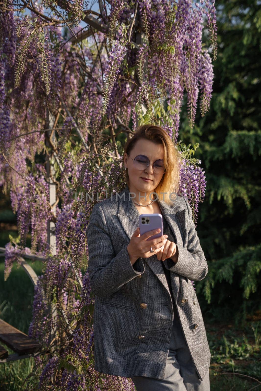 A woman in a grey blazer and skirt stands in front of a blooming wisteria tree, looking at her phone.