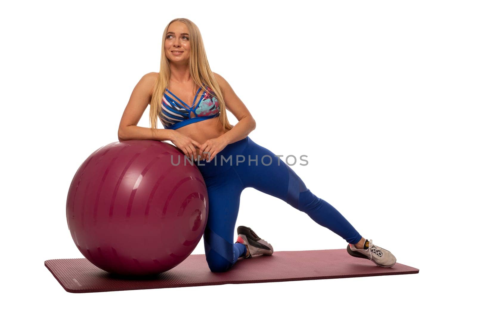 Full body of young fit female in sportswear sitting on fitness ball and looking at camera while resting during workout against white background