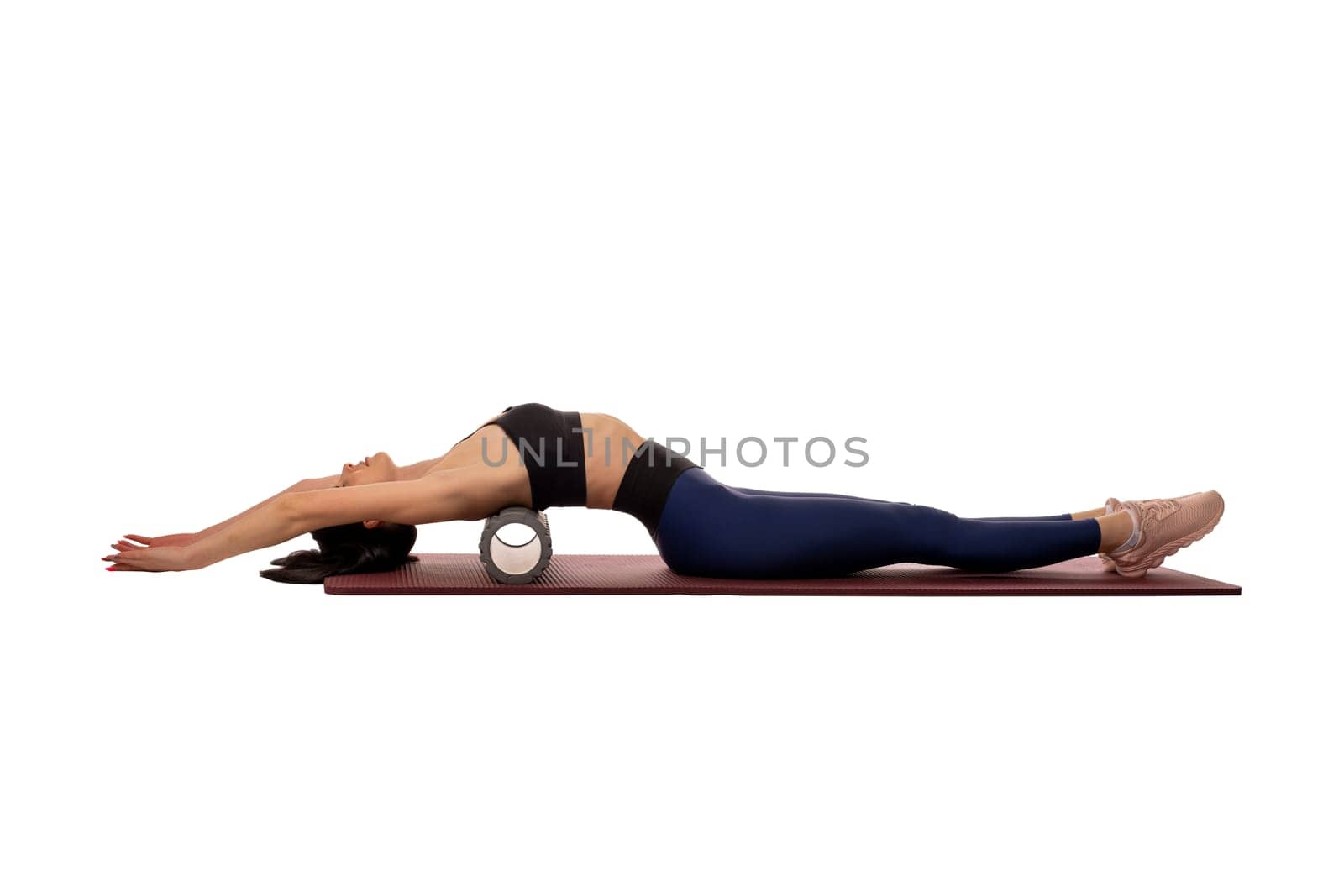 Side view of graceful young female athlete with long dark hair in activewear lying on mat with foam roll behind back isolated