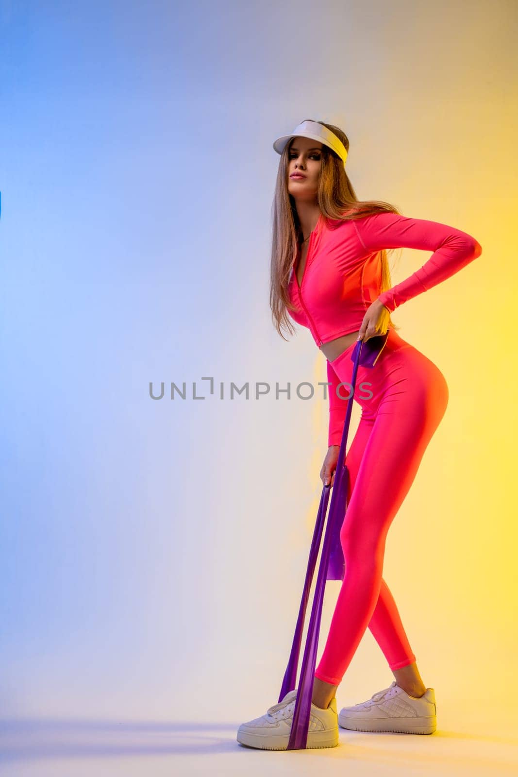 Full body side view of confident young female in sportswear and cap standing with hands on waist and looking at camera against blue background