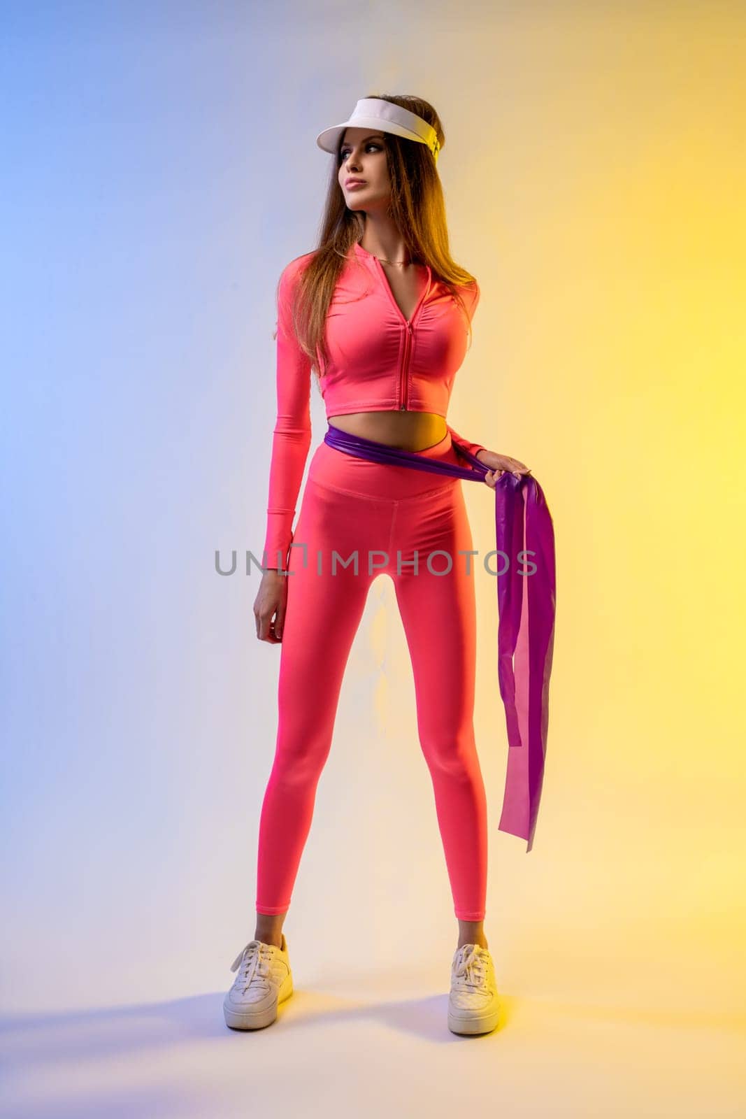 Full body of confident young fit female athlete with long brown hair in stylish pink sportswear and cap standing with resistance band in light studio