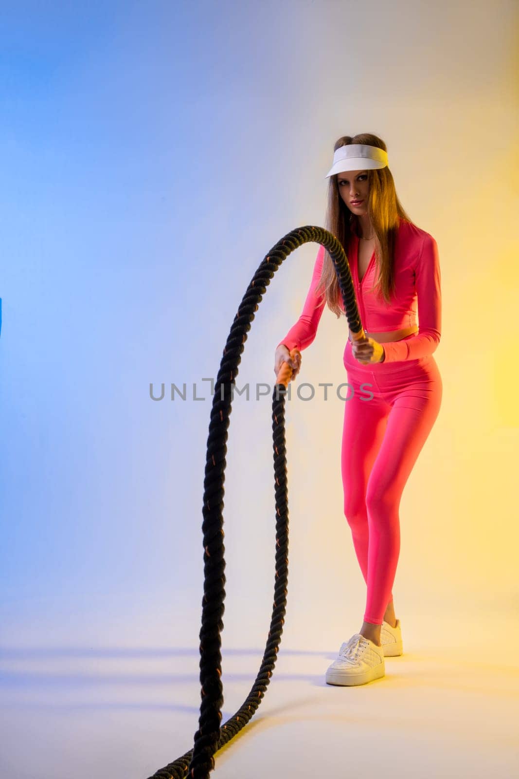 Full body of serious young female model with long hair in stylish activewear standing in bright color studio with battle rope