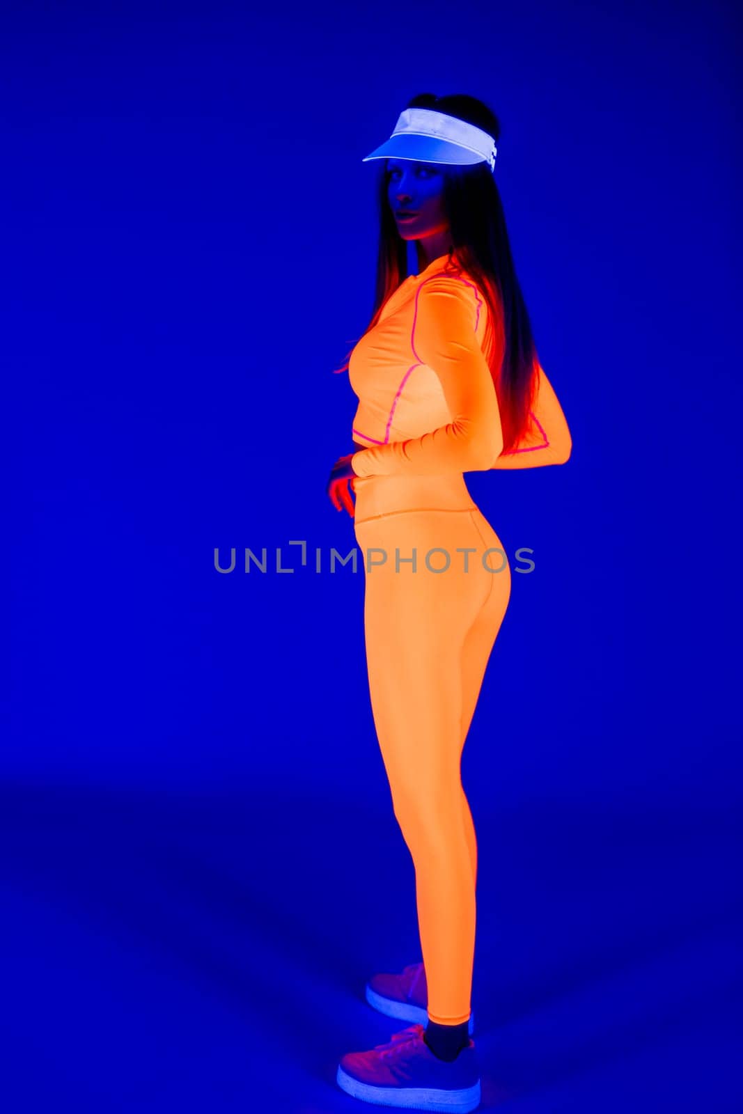 Full body side view of slim young female model in trendy outfit and cap standing against blue background in studio