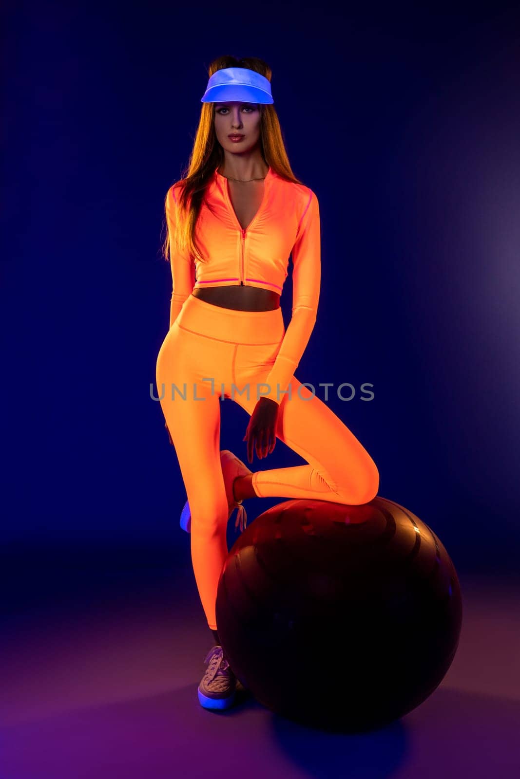 self assured young female athlete with long hair in stylish bright neon orange sportswear and blue cap leaning knee on fit ball