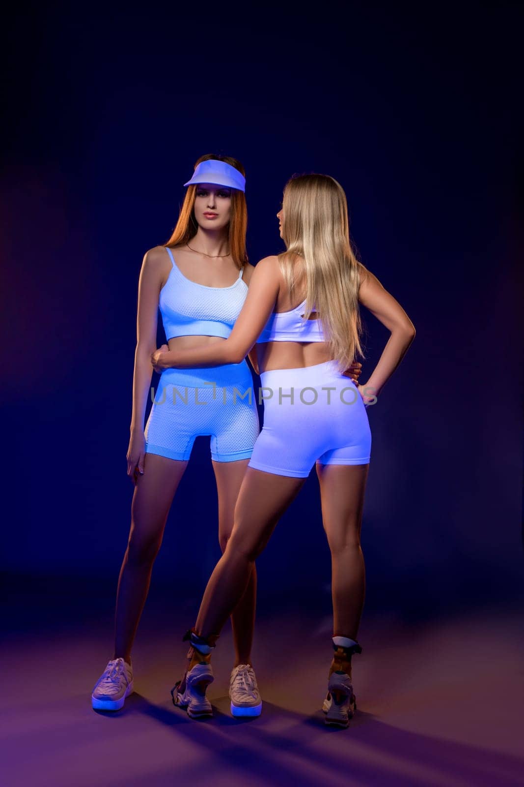 Full length of confident young fit female models with long brown and blond hair in white sportswear standing together in blue studio