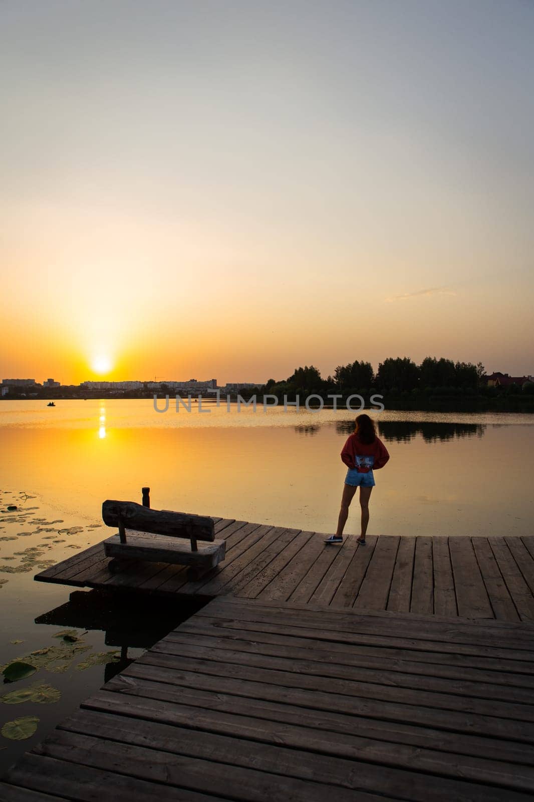 A scenic lakeside view at sunset with a woman standing on a wooden pier, featuring a wooden bench and golden reflections on calm water.