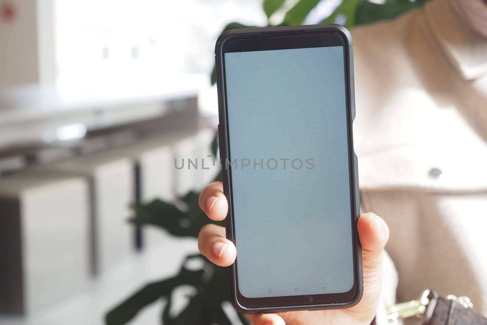 A person shows a blank smartphone screen while indoors surrounded by greenery.