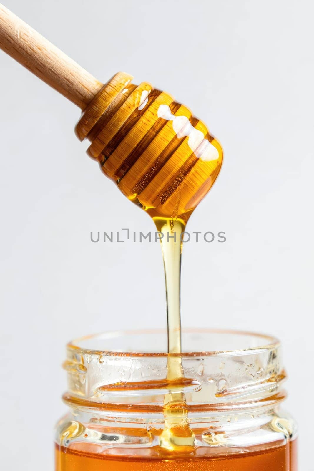 Golden honey elegantly drips from a wooden dipper into a clear glass jar. The bright background enhances the natural sweetness, showcasing the rich texture and color of the honey.