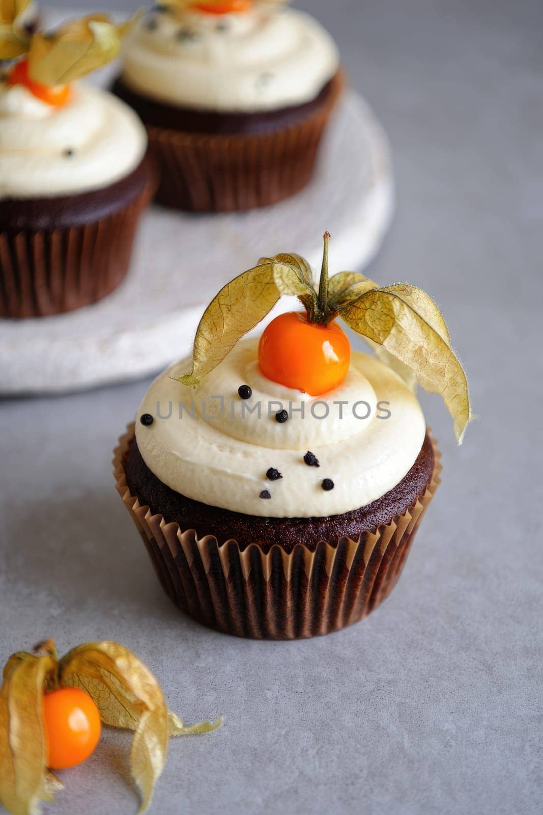 Chocolate cupcakes feature creamy white frosting and a bright fruit topping, accompanied by a delicate golden husk. The rustic gray surface enhances the inviting appearance of the treats.