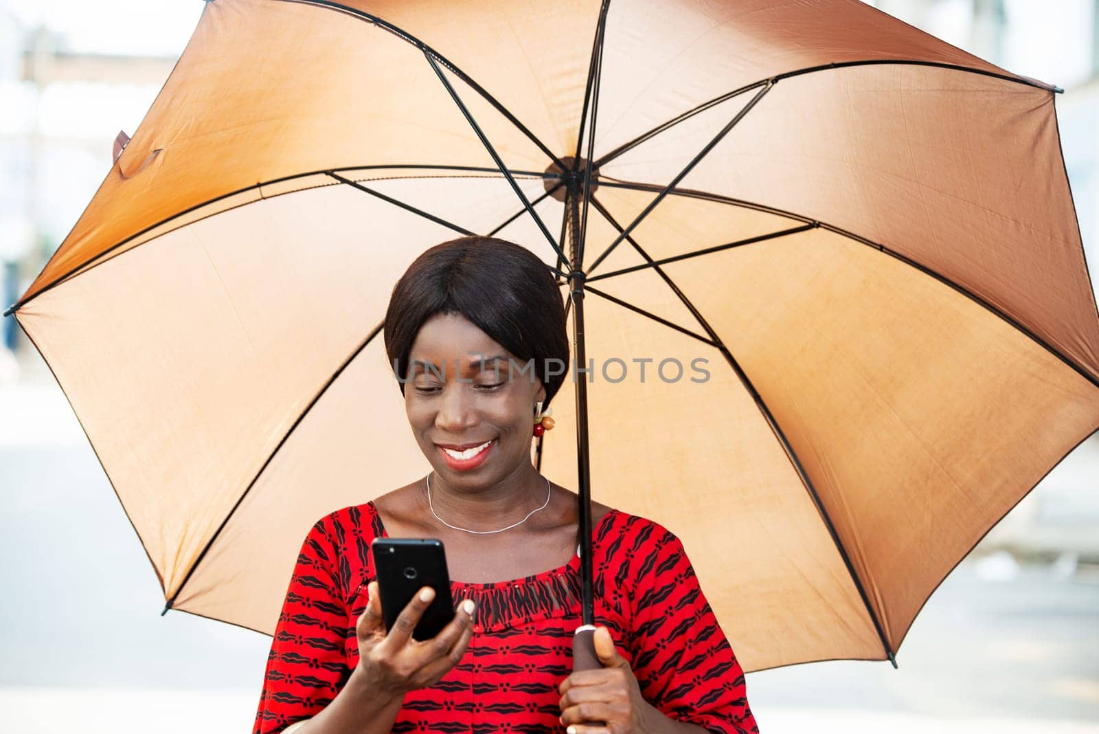 mature woman in traditional clothes standing looking at mobile phone smiling with umbrella over her head.