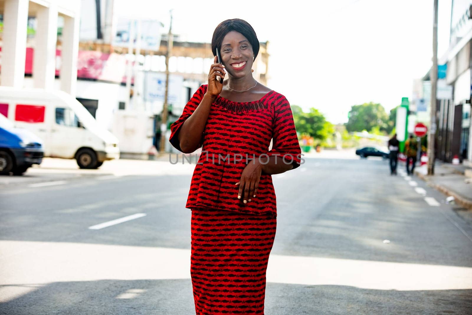Happy african adult business woman, dressed in red loincloth, is communicating on the phone while walking in the street