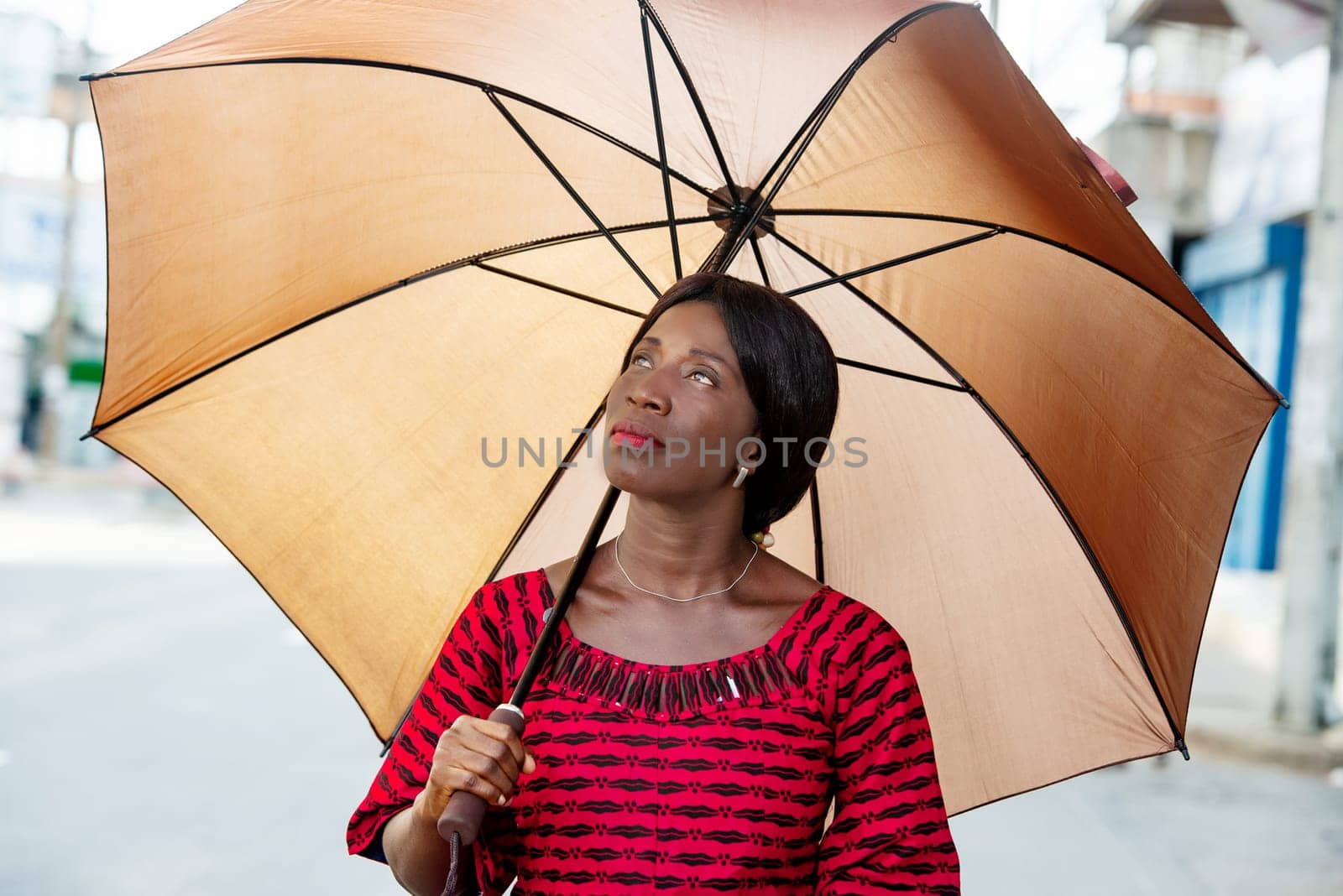 beautiful african adult businesswoman dressed in red loincloth holding umbrella in hand and looking up smiling in the street