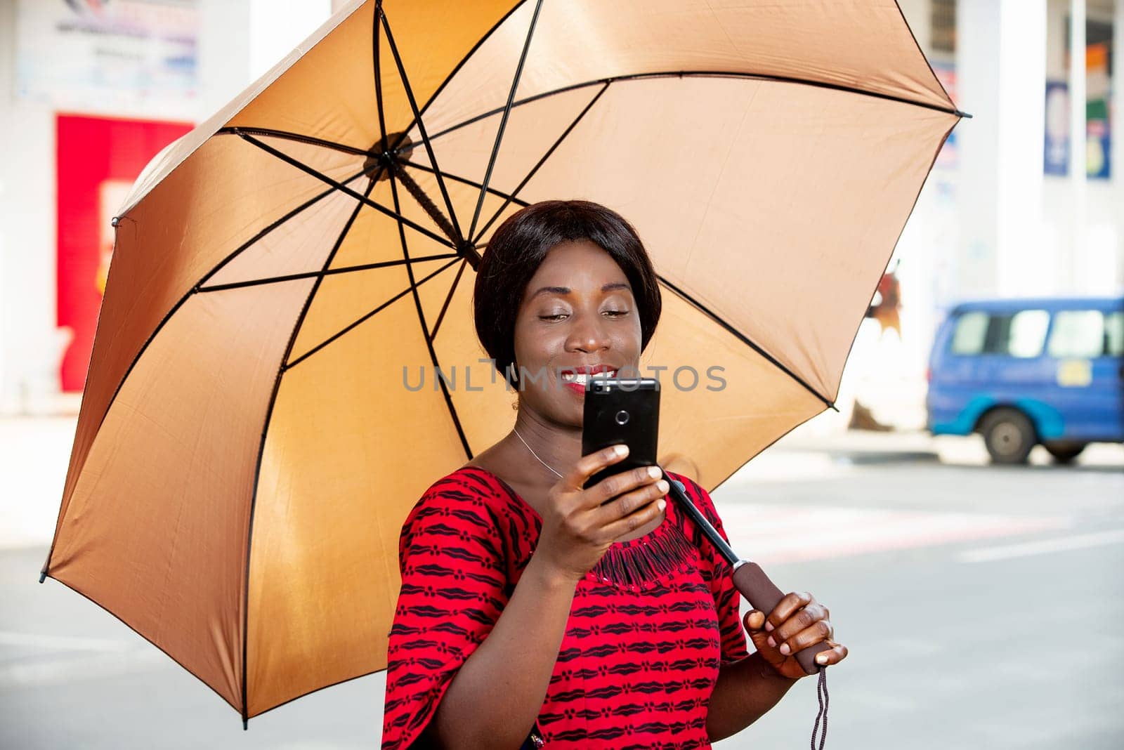 beautiful african adult businesswoman standing, holding an umbrella in hand, taking a self portrait photo in the street or chatting on video on her mobile phone smiling