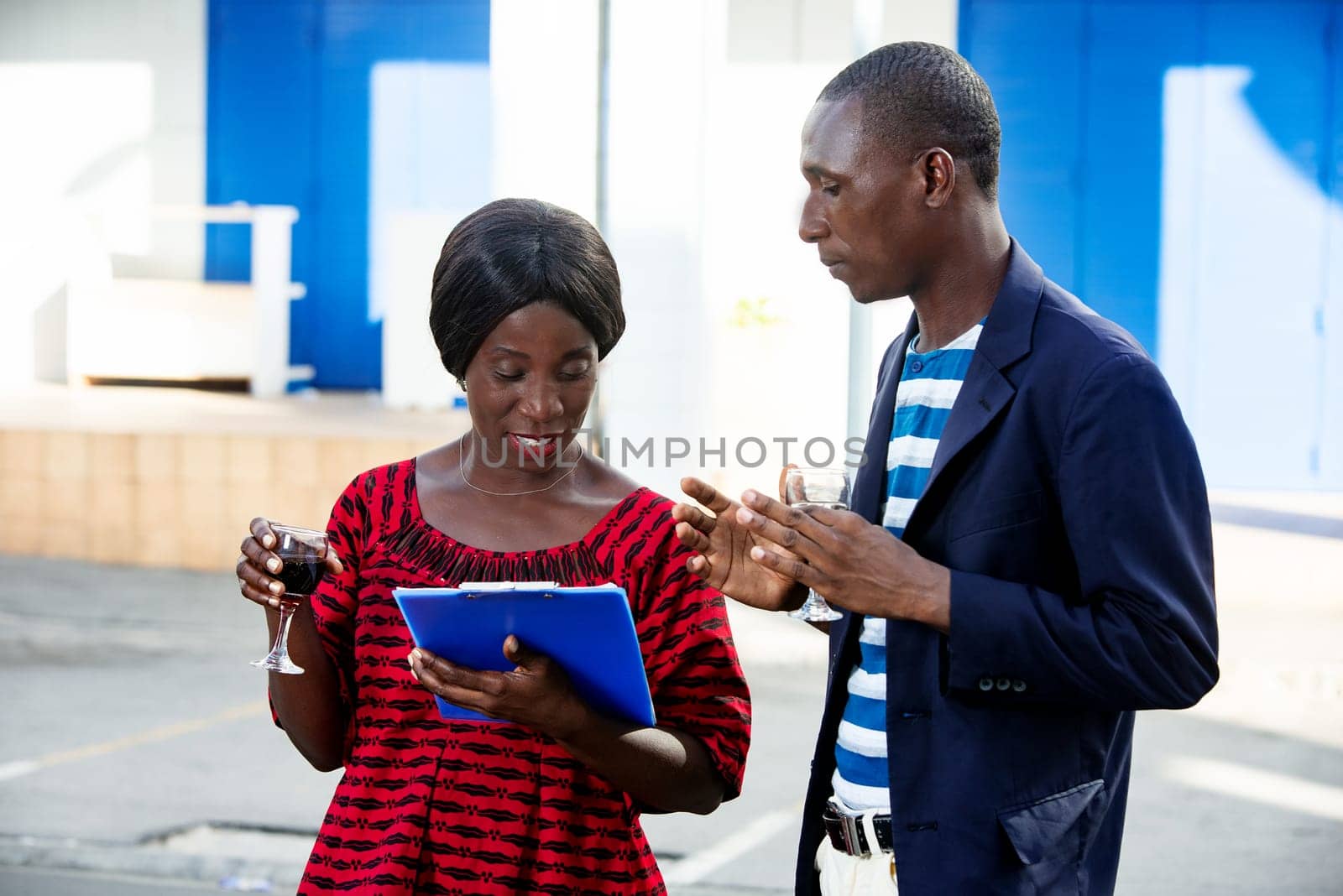 handsome man and beautiful african adult businesswoman standing by discussing project business, holding in hand a blue document and a drink, on the street