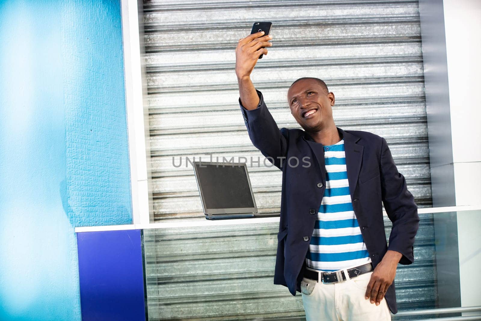 Handsome african adult man making selfie photo on cellphone camera while standing in city street in sunny summer day. Smiling man having an online video call on smartphone during free time outdoors