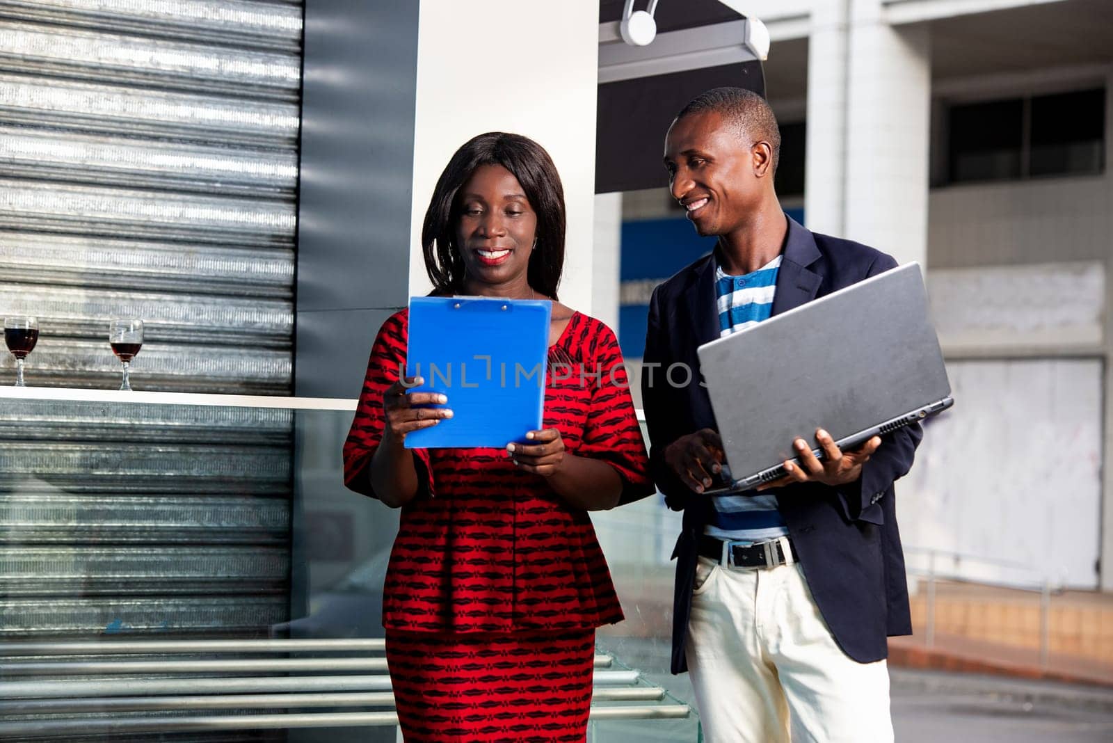 businessman holding a computer chatting with businesswoman holding the folder with documents in hand outdoors smiling together