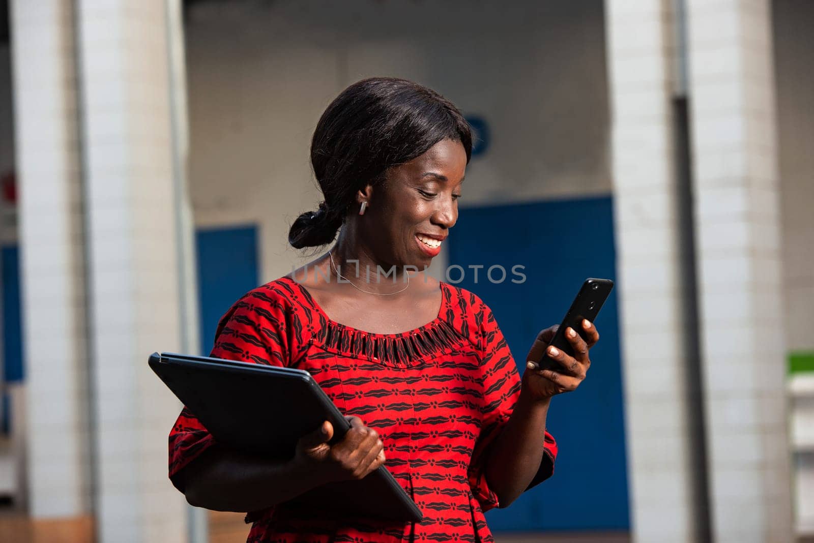beautiful african businesswoman dressed in red loincloth standing in the street is looking at her phone holding a laptop in hand