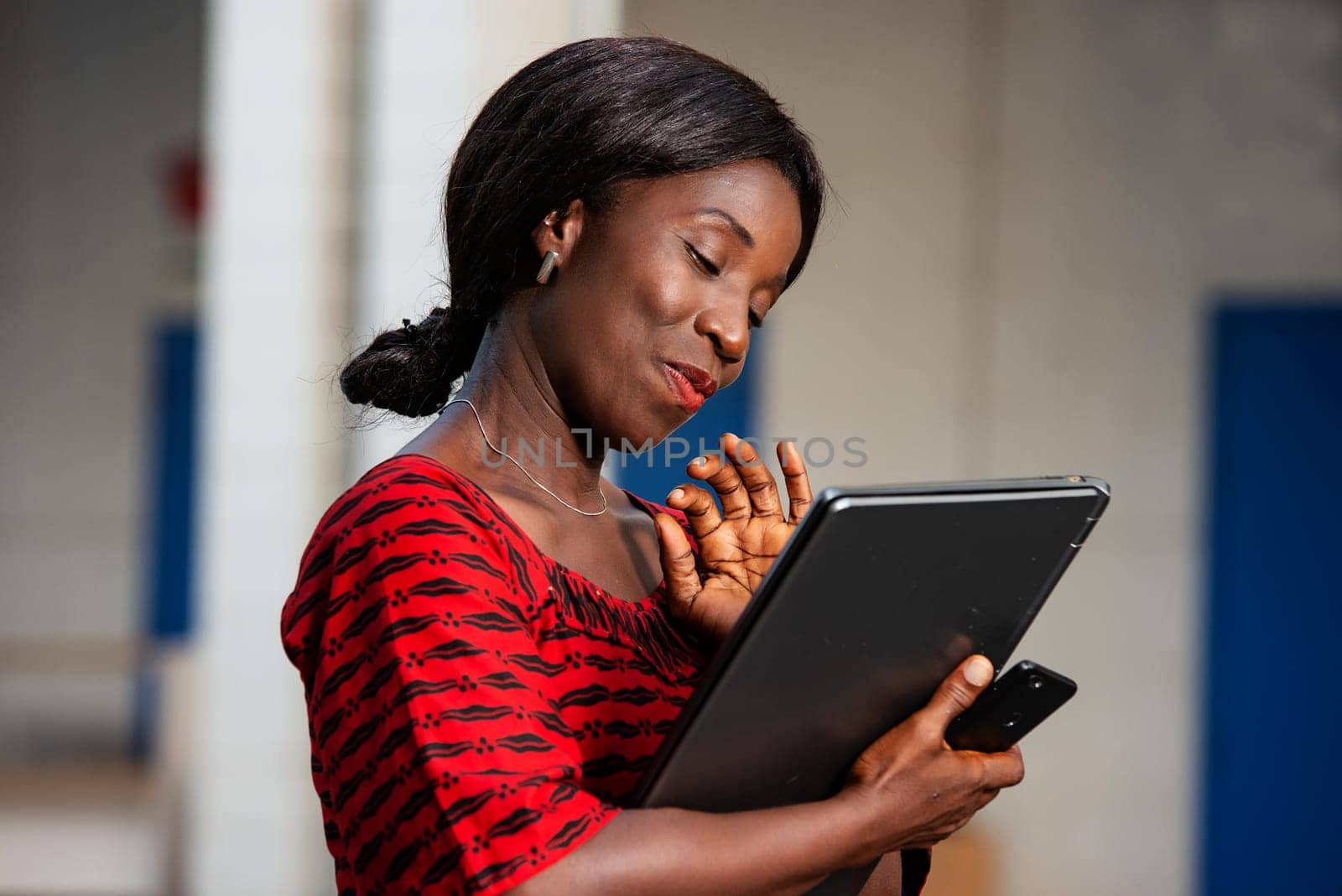 beautiful African businesswoman dressed in red loincloth standing on the street holding in hand a laptop and phone