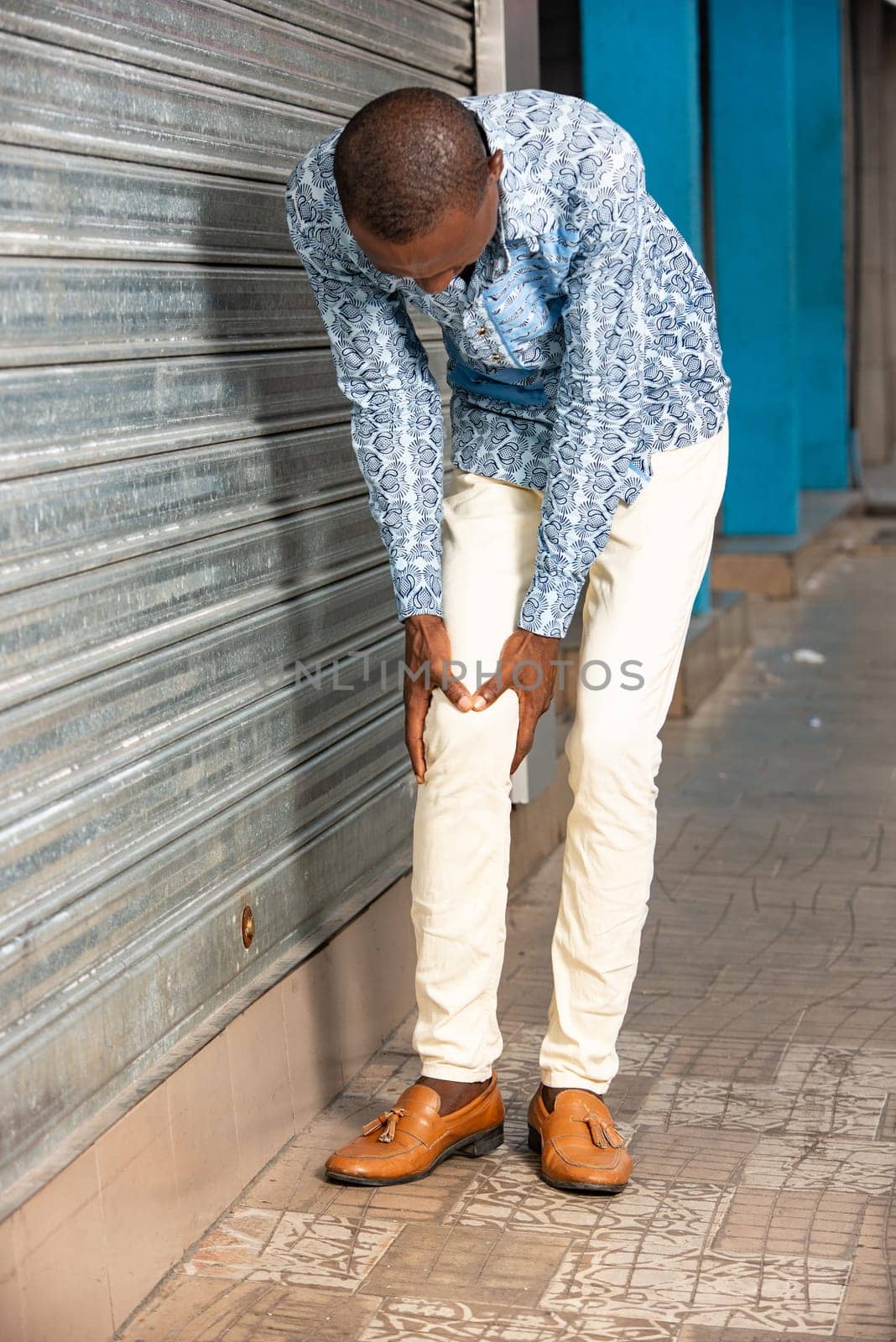young african businessman holding knee with his hands in pain after suffering a muscle injury while walking down the street