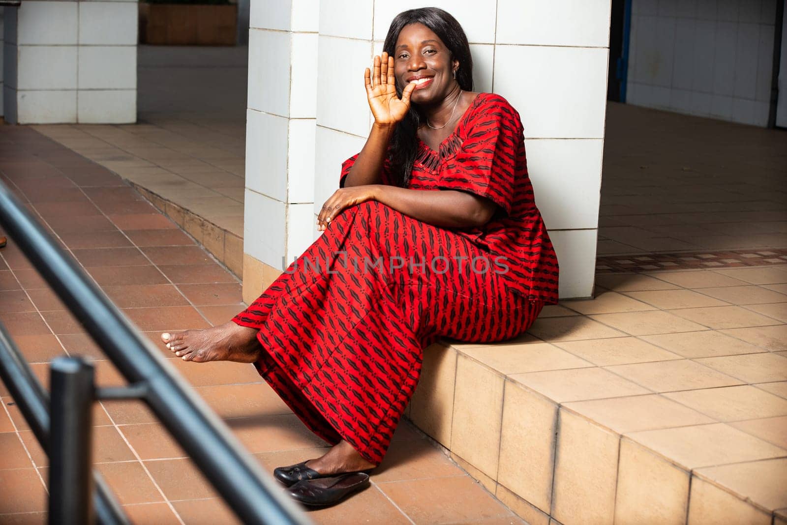 beautiful African woman in traditional dress sitting near a wall smiling at the camera while making hand gesture.