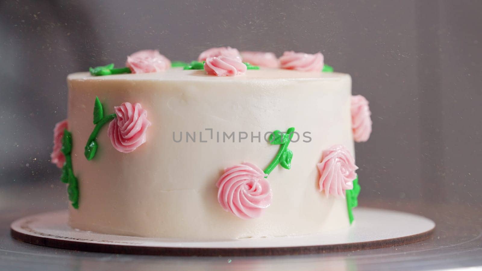 Beautiful buttercream cake on turntable. Decorated with delicate pink flowers, green leaves on smooth white icing. Bakery, celebration, wedding, pastry concept. High quality