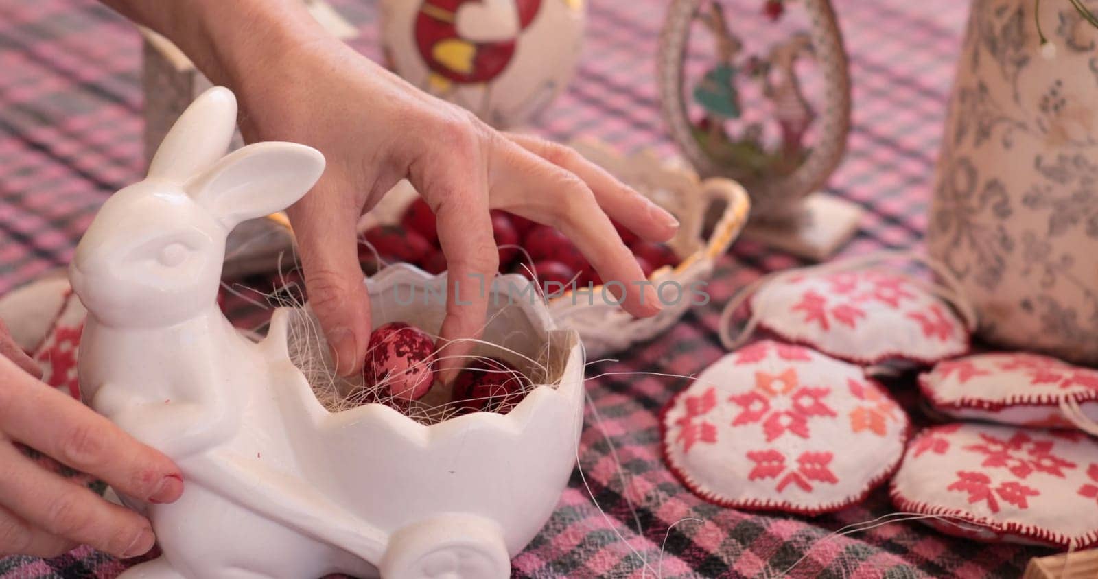 Hands place a red Easter egg into a ceramic holder, surrounded by decorations on a checkered tablecloth. Coloring eggs creates joyful moments during the Easter celebration.
