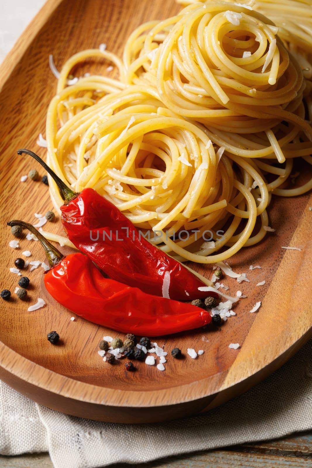 Twirls of pasta sit elegantly on a wooden plate, accompanied by two vibrant red chili peppers and a sprinkle of coarse salt and black pepper, creating a tempting presentation.