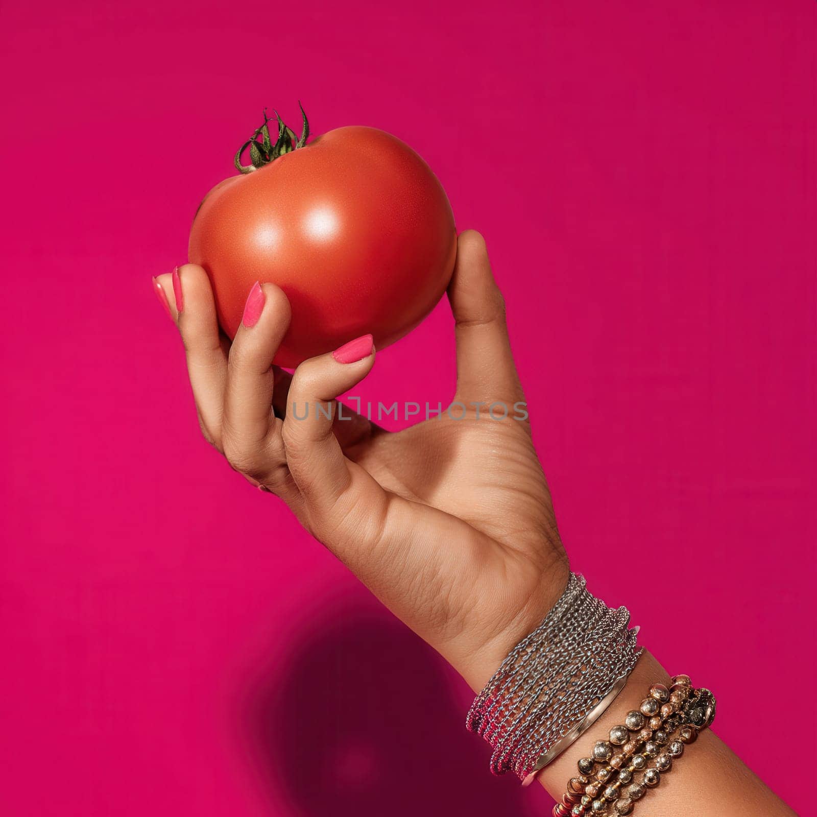 A hand with bracelets and pink manicure holding a ripe tomato against a vivid pink background. Ideal for food styling, pop-art compositions, beauty branding, and bold lifestyle campaigns