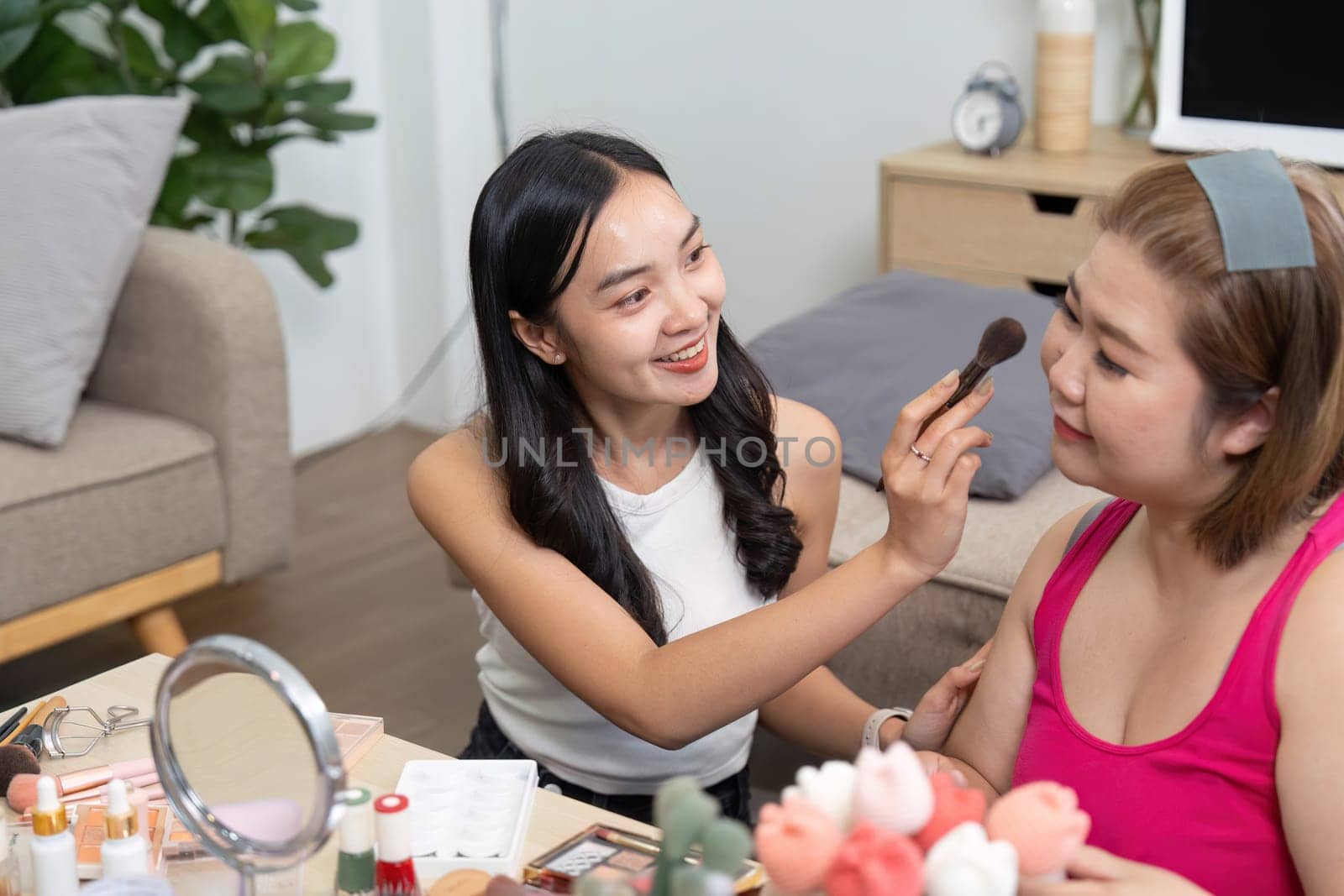 Two friends share a joyful moment as one applies makeup on the other, demonstrating creativity and connection in a cozy environment.