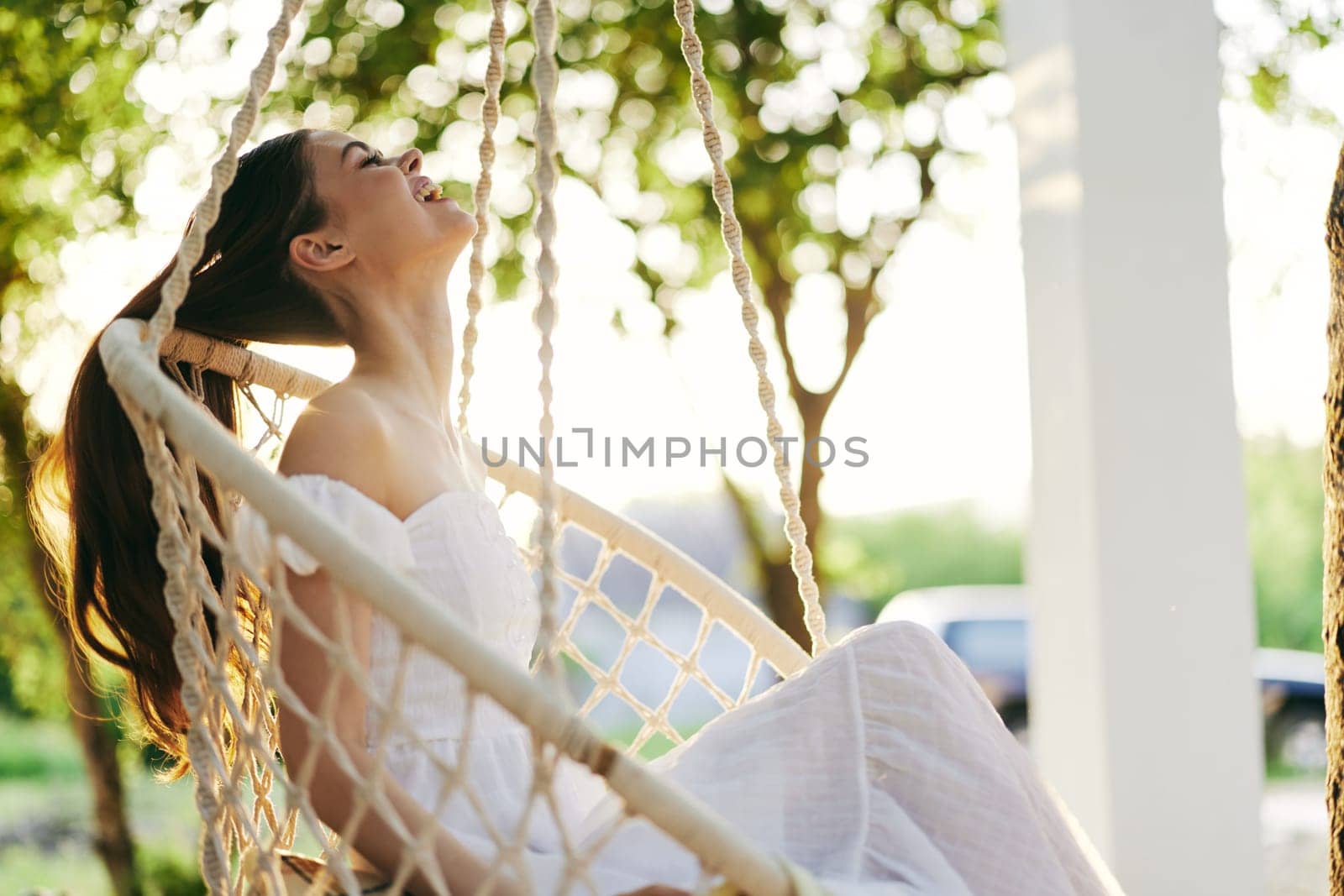 A joyful young woman in a flowing white dress sits on a swinging chair, surrounded by lush greenery and sunlight, embodying peace, happiness, and relaxation in nature