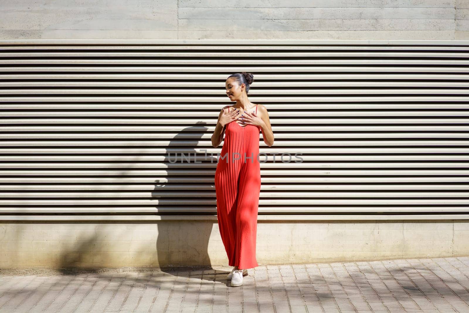 A stylish and fashionable woman poses confidently in a vibrant and eyecatching red dress, standing proudly against a modern textured wall, showcasing her contemporary style and elegant personality