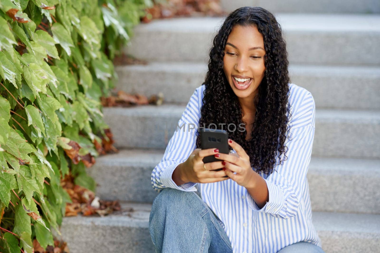 A cheerful young woman with curly hair is seated on steps, smiling as she uses her smartphone. Lush green ivy frames the scene, creating a vibrant atmosphere of warmth and happiness