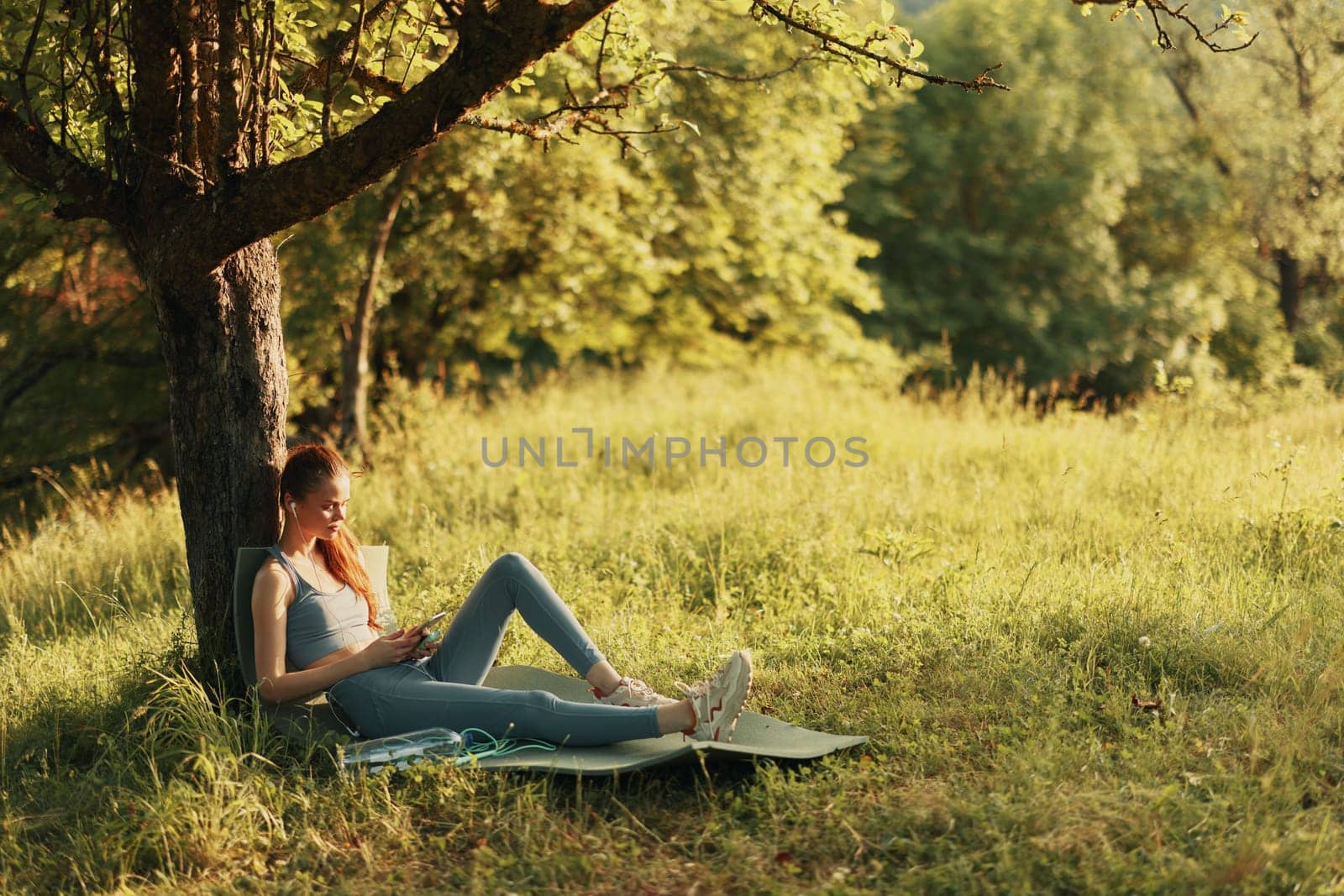 A serene young woman in comfortable fitness attire reclining against a tree, embodying relaxation and wellness in a lush green setting, ideal for promoting a healthy lifestyle or outdoor activities