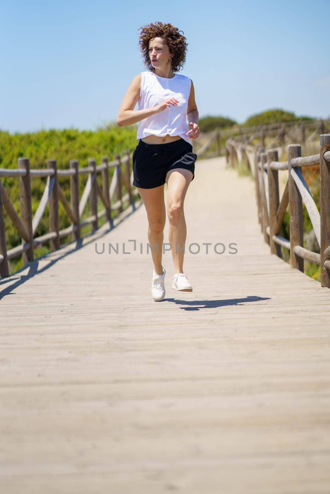 Full body of active female athlete in sportswear and sneakers running along wooden boardwalk during fitness training