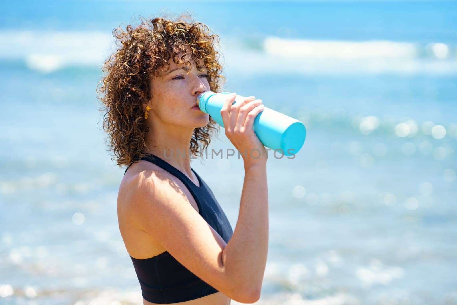 Side view of young fit sportswoman with curly hair drinking fresh water after training near waving sea on sunny day