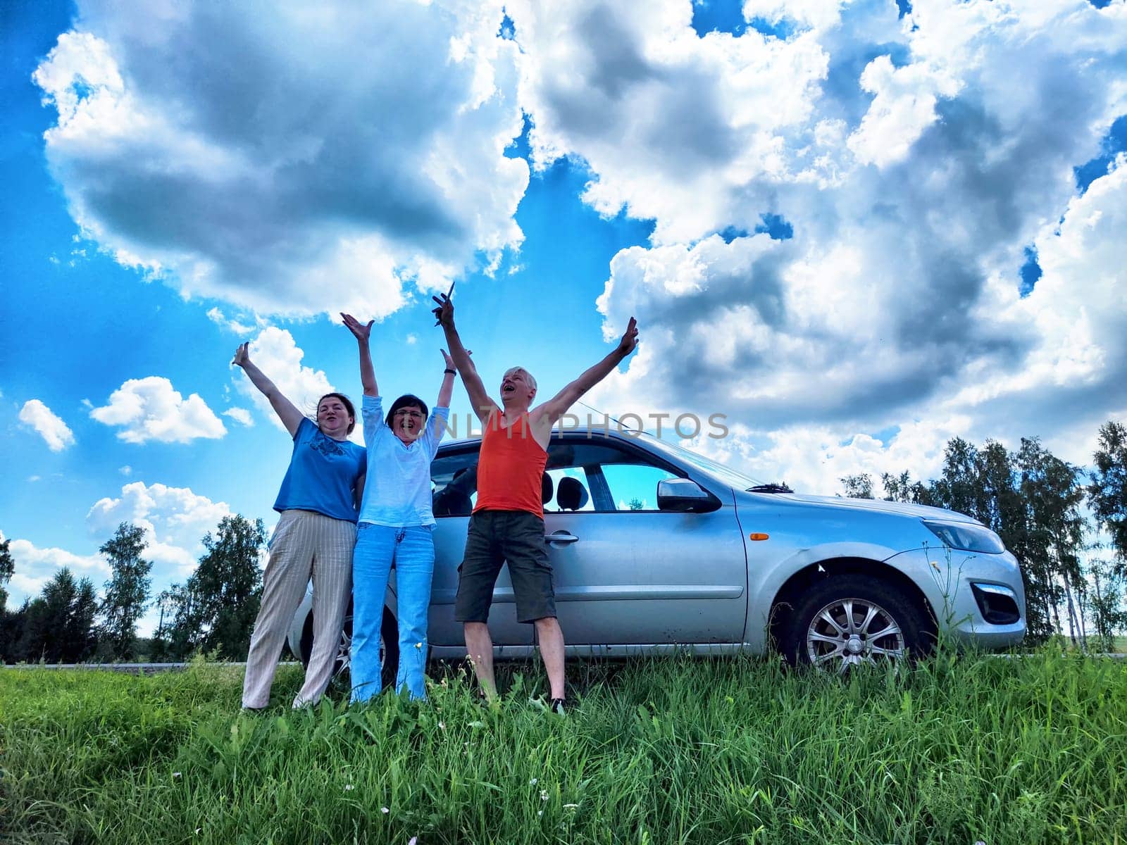A happy couple stands with arms raised by their car, celebrating their travel adventure under a bright blue sky with fluffy clouds. They showcase joy and togetherness.