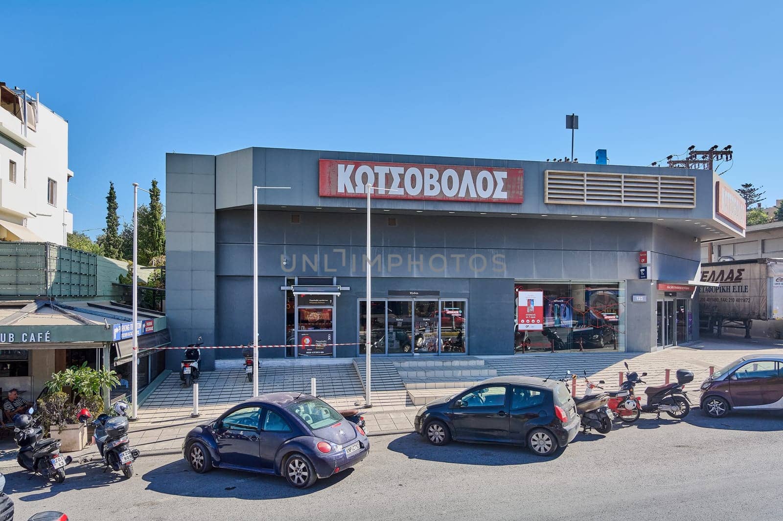 Crete.Greece - april 13, 2025: A busy urban street corner in Heraklion features a retail store named , a caf with outdoor seating CLUB CAFE, and other local businesses. Parked motorcycles.