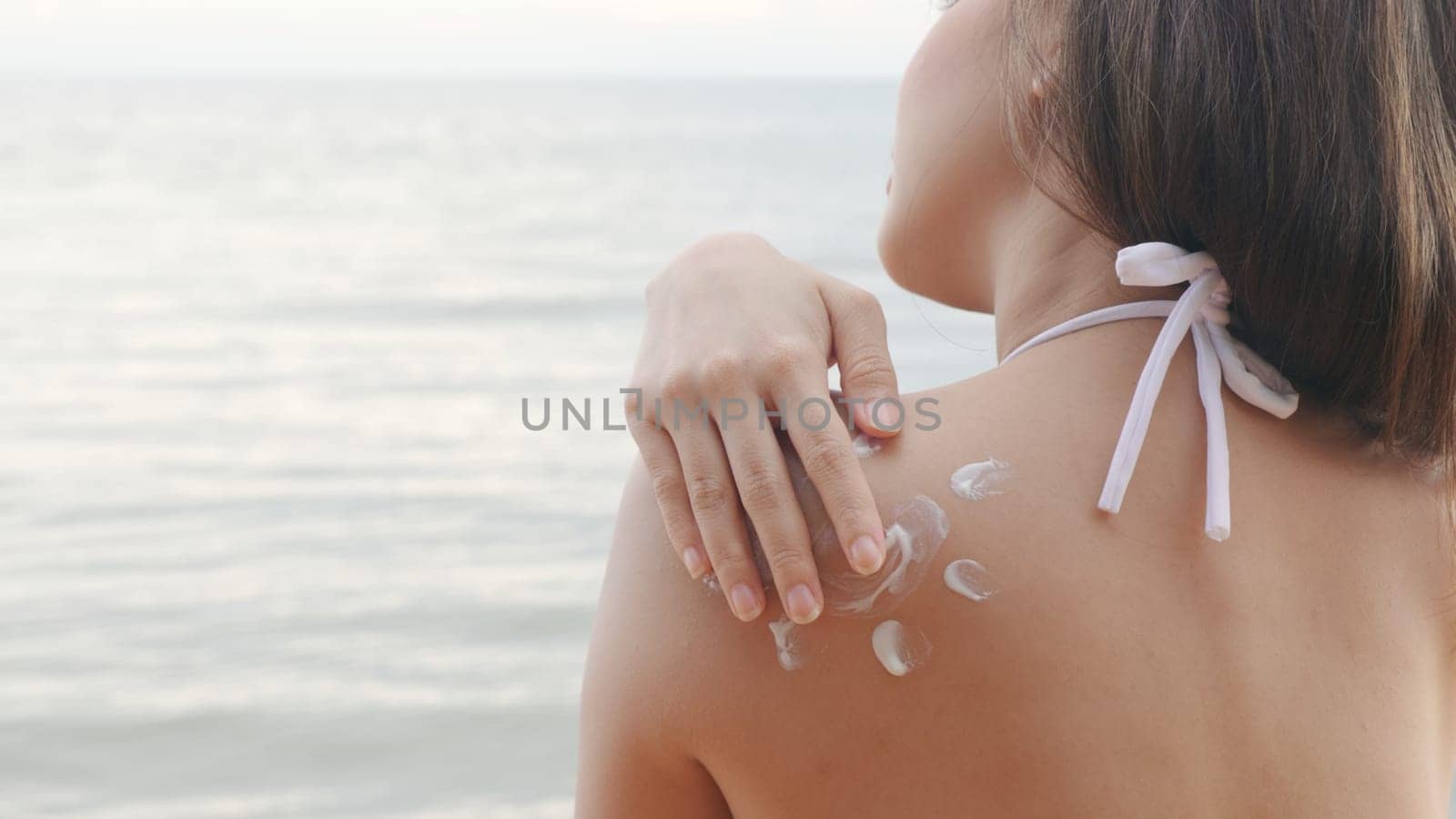 Beautiful Asian woman applying sun protection cream on tanned shoulder on the beach, sun Cream, Close up female in bikini using sunscreen lotion on back body before sunbathing