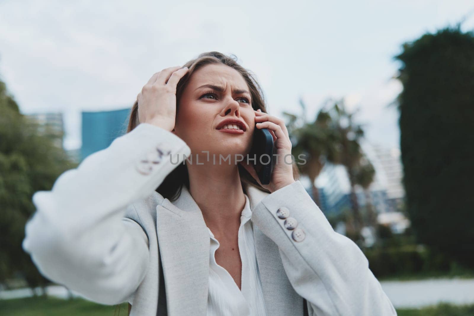 A worried businesswoman dressed in a stylish gray blazer communicates on the phone in an urban outdoor environment. The scene captures the emotional tension and stress of a modern professional during the day.