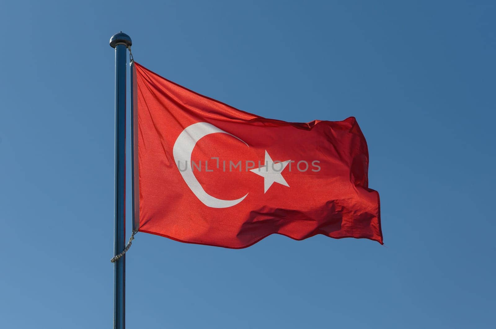 A vibrant red flag adorned with a white crescent and star flutters proudly in the gentle breeze. The clear sky enhances the bold colors, creating a striking contrast on a beautiful day.