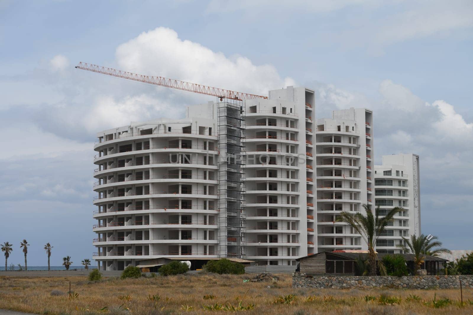 Amidst a tranquil landscape, a modern residential structure rises towards the sky, partially completed. The coastline glimmers in the background under a moody, cloudy sky.