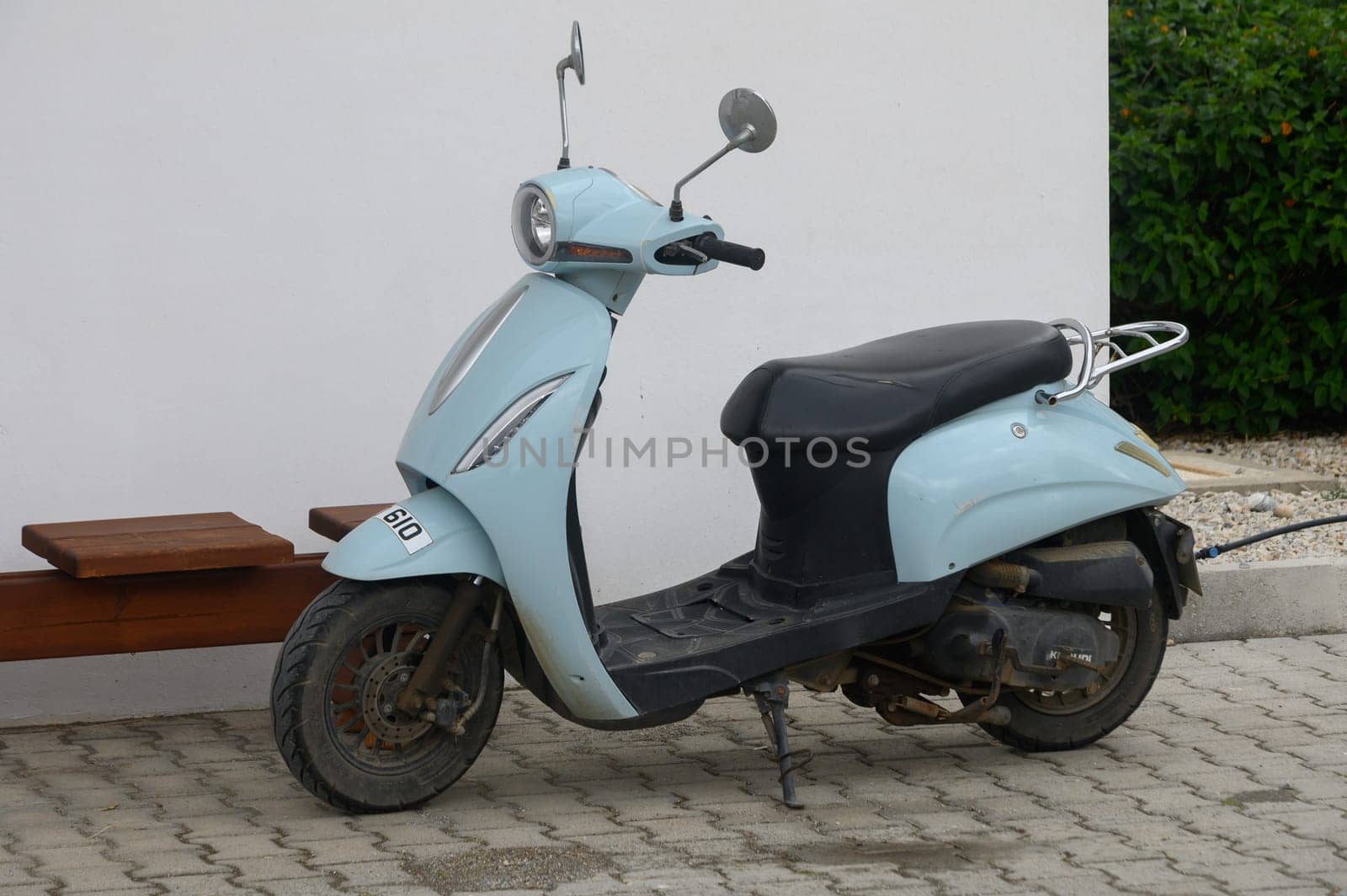 Gaziveren Cyprus -10.04.2025 A charming blue scooter sits quietly beside a pristine white wall, surrounded by lush green foliage, capturing a moment of stillness on a bright day.