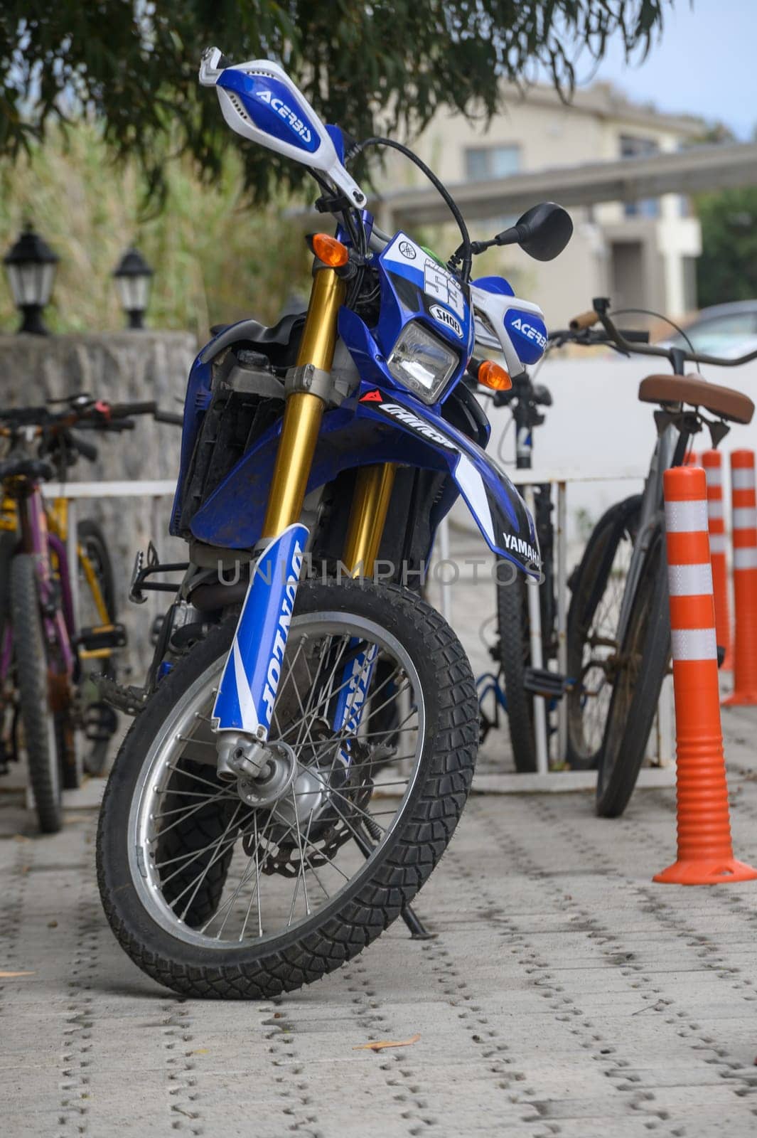 Gaziveren Cyprus -10.04.2025 A vibrant motorcycle stands parked next to bicycles, surrounded by lush greenery and decorative lamps. The setting exudes an active urban lifestyle, enhanced by soft sunlight.